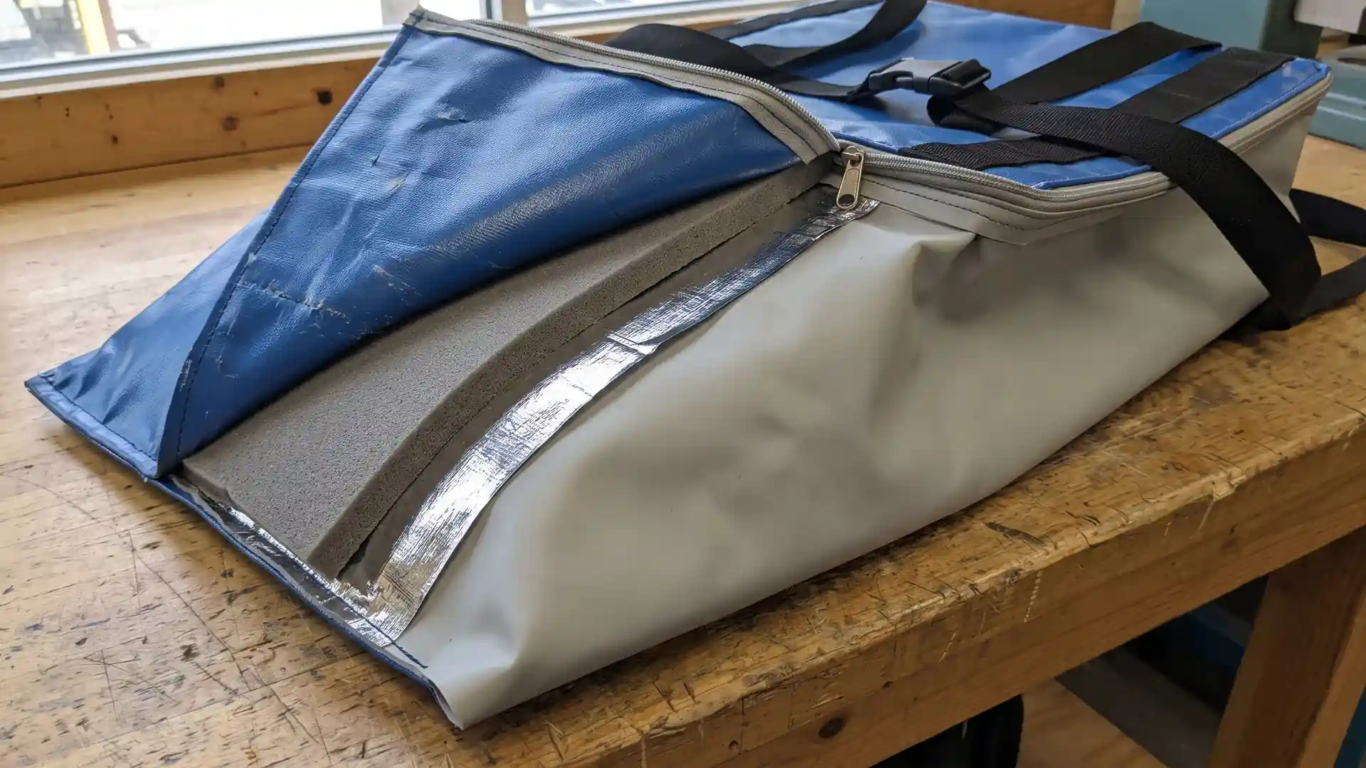 Peeled-back wall corner of a reusable cold chain delivery bag showing tarpaulin shell, closed-cell foam, radiant barrier, and smooth TPU liner.