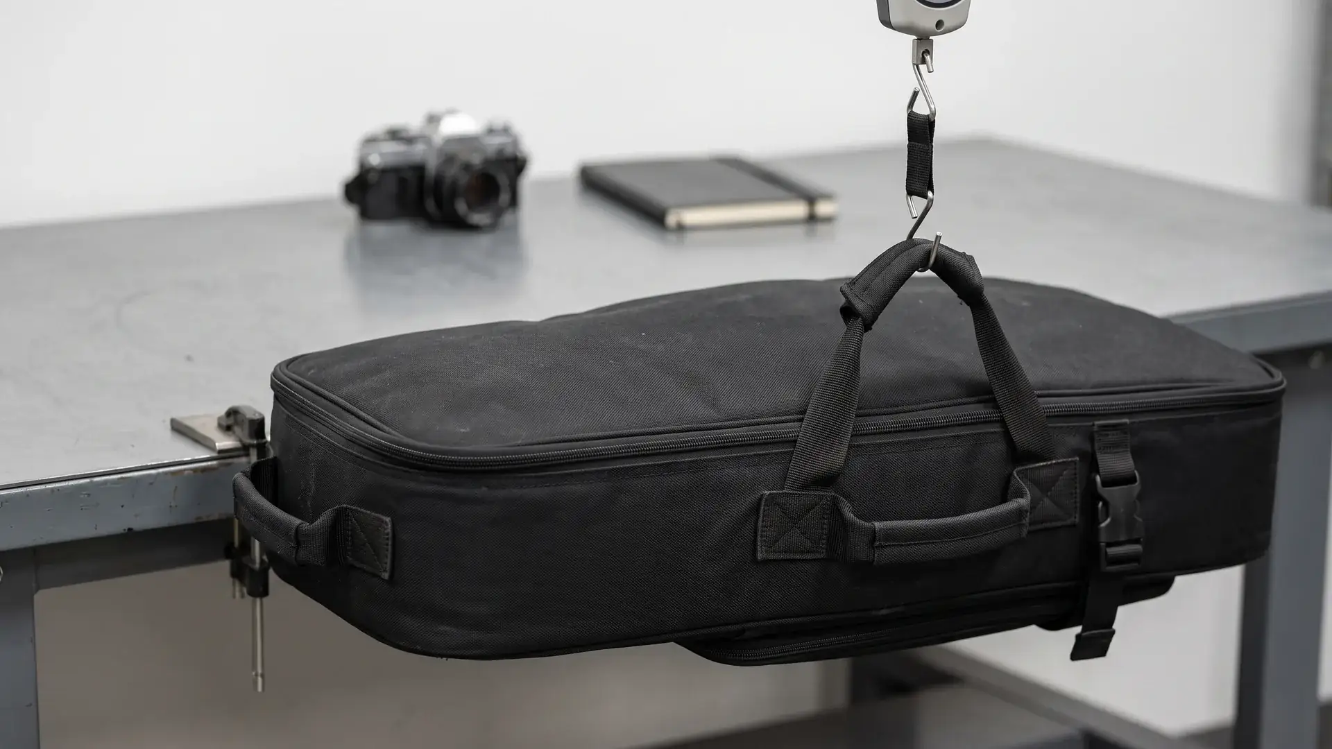 Loaded instrument carrying case closed on a QC worktable with a mechanical tension gauge pulling one handle straight upward while the opposite handle is held flat against the table edge.