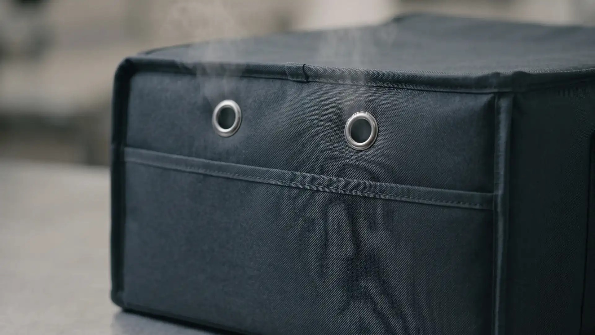 Upper side panel of a commercial pizza delivery bag with metal steam-vent grommets releasing a faint haze to prevent soggy crusts.